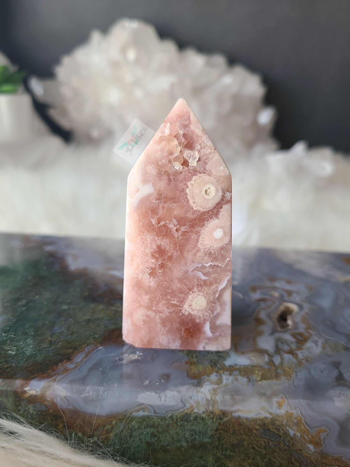 A pointed pink stone tower or crystal point with natural white and peach marbling, standing upright on a polished agate slab with blurred white crystals in the background.